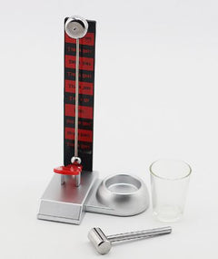 Compact drinking game setup featuring a small silver hammer, a clear shot glass, and a vertical scoreboard with red and black labels, designed for lively party fun.