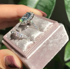 Close-up of a geometric-patterned ring featuring an oval blue zircon gemstone surrounded by sparkling clear stones, displayed in a soft pink velvet box.