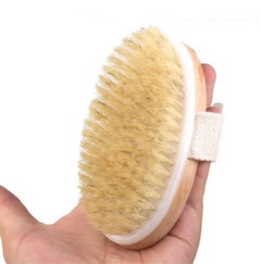 Hand holding a natural bristle bath brush with a wooden base and fabric strap designed for exfoliating and cleansing skin.