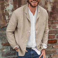 Man wearing a beige lightweight cotton linen blazer over a white casual shirt, standing against a rustic brick and metal wall, smiling with one hand in pocket.