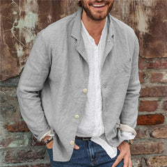 Man wearing a light gray slim fit casual cotton linen blazer over a white shirt, standing against a rustic brick and weathered wall background, smiling with hands in pockets.