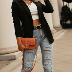 Woman wearing a slim black long-sleeve blazer over a cropped white top paired with distressed light blue jeans, accessorized with layered necklaces and a tan handbag.