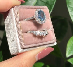 A hand holding a soft pink velvet ring box displaying two geometric-patterned rings, one featuring a sparkling blue zircon center stone surrounded by smaller clear stones.