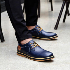 Men's navy blue genuine leather lace-up casual dress shoes with contrasting tan laces and stitching, paired with black pants on a smooth floor.