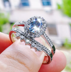 Close-up of a silver geometric-patterned ring featuring a sparkling oval blue zircon surrounded by smaller clear stones, held between fingers with a blurred outdoor background.