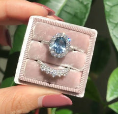 A hand holding a pink velvet ring box containing a geometric-patterned blue zircon ring surrounded by small clear stones, paired with a silver curved band encrusted with diamonds.