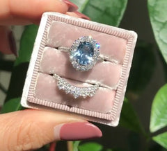 Hand holding a small velvet ring box containing a geometric-patterned silver ring with a sparkling oval blue zircon center stone and a matching diamond-accented band.