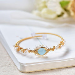 Delicate gold wire cuff bracelet featuring a braided design with small white pearls and a central light blue gemstone, styled on an open book with soft floral background.