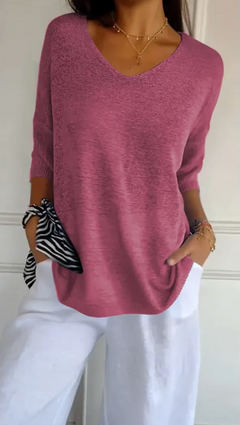 Woman wearing a stretchable, block-colored V-neck knit sweater in muted pink paired with white pants, accessorized with layered gold necklaces and bracelets, holding a zebra print scarf.