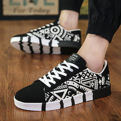 Black low-top sneakers featuring bold white geometric graffiti patterns, paired with white laces and a unique striped sole, worn with casual black pants on wooden floor.