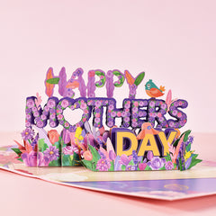 Pop-up floral Mother's Day card with colorful purple flowers, green leaves, a small bird, and the words "Happy Mothers Day" in vibrant, decorative letters on a pink background.