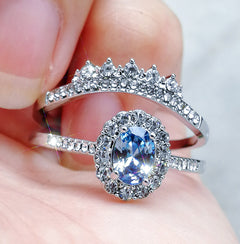 Close-up of a silver geometric ring featuring a sparkling oval blue zircon center stone surrounded by smaller clear stones, held between fingers against a neutral background.