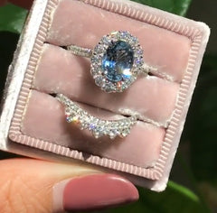 Two delicate rings displayed in a plush pink velvet box, featuring an oval blue zircon center stone surrounded by a geometric halo and a curved band with sparkling accents.