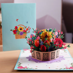 Intricately designed 3D pop-up flower bouquet card featuring colorful roses, sunflowers, and greenery in a decorative basket, set inside a pastel blue greeting card.