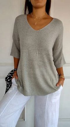 Woman wearing a stretchable light gray V-neck knit sweater with three-quarter sleeves paired with white pants, accessorized with layered gold necklaces and bracelets.