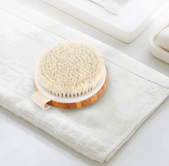Round bath brush with soft natural bristles and wooden handle resting on a folded white towel in a bathroom setting for gentle exfoliation and skin care.