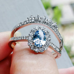 Close-up of a silver ring featuring a geometric design with a sparkling oval blue zircon center stone surrounded by small clear stones, worn on a finger outdoors.