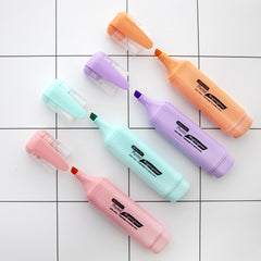 Three pastel-colored highlighters in peach, lavender, and mint green with pocket clips are arranged on a white grid surface, each with its cap removed to show the chisel tips.