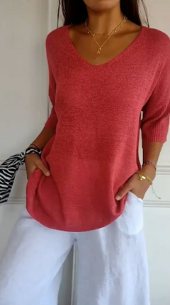 Woman wearing a loose-fitting red v-neck knit sweater with three-quarter sleeves, paired with white pants, accessorized with layered gold necklaces and bracelets, holding a patterned scarf.
