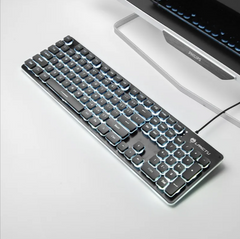Black USB wired membrane keyboard with 104 backlit keys and luminous edges, connected to a computer monitor on a white desk.