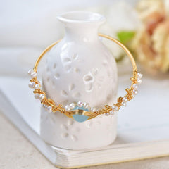 Elegant gold wire braided bracelet adorned with small baroque pearls and a central light blue stone, styled vintage fashion accessory displayed on white ceramic vase.