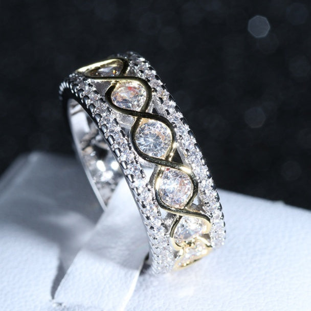 Close-up of a two-tone gold and silver ring with round cut cubic zirconia stones set in a wide, multi-row infinity design on a white display stand against a dark background.