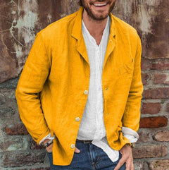 Man wearing a bright yellow slim fit cotton linen blazer over a white shirt and blue jeans, standing against a rustic brick and weathered metal wall.