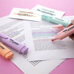 Pastel-colored highlighters with chisel tips highlighting and underlining study cards and text on white paper, set against a pink background.