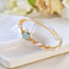 Delicate gold wire braided cuff bracelet featuring a central light blue stone and accented with small white pearls, styled on an open book with soft floral background.