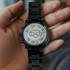 Hand holding a black metal watch with an engraved back featuring an intertwined heart design and the phrase "I will love you until the end of time" as a sentimental gift.