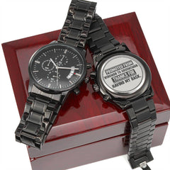 Two black men's chronograph watches with metal bands displayed on a polished wooden box, one showing a classic black dial and the other engraved with a humorous groomsman message on the back.