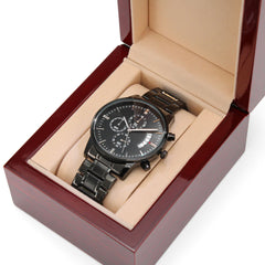 Black men's chronograph watch with engraved detail, featuring a metal link band, displayed in a polished wooden gift box with a beige cushion interior.