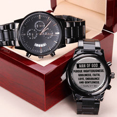 Two men's black chronograph watches with metal bands displayed on and beside a polished wooden gift box, one showing a sleek black dial and the other the engraved caseback with a faith-inspired scripture message.