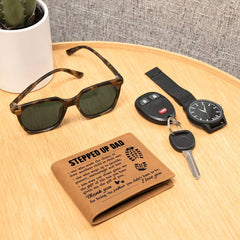 Leather bifold wallet with heartfelt "Stepped Up Dad" message, placed on a wooden surface alongside sunglasses, a wristwatch, and car keys.