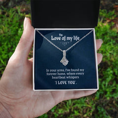 Hand holding an open jewelry box displaying a silver necklace with a delicate ribbon-shaped pendant and a card reading "To Love of my life" with a romantic message inside, set against an outdoor green background.