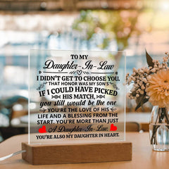 Clear acrylic plaque with black and red heartfelt message for a daughter-in-law, illuminated by a light-up wooden base, displayed on a table next to a vase of flowers.