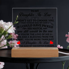Clear acrylic plaque with a heartfelt message to a daughter-in-law, illuminated by a light-up wooden base, surrounded by soft pink flowers on a dark surface.