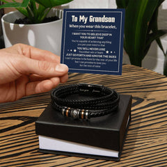 Hand holding a card with a heartfelt message for a grandson above a braided black leather bracelet with engraved metal accents resting on a black and white gift box.