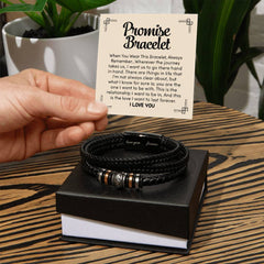 Hand holding a card with a heartfelt message next to a black braided leather bracelet with metal accents, displayed on a black gift box on a wooden surface with green plants in the background.