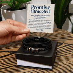 Hand holding a card with a sentimental message above a black braided men's bracelet with metal accents, displayed on a black gift box on a wooden surface with potted plants in the background.