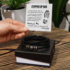 Hand holding a heartfelt message card for a stepdad next to a black braided leather bracelet with engraved metal accents resting on a black gift box, with green plants in the background.
