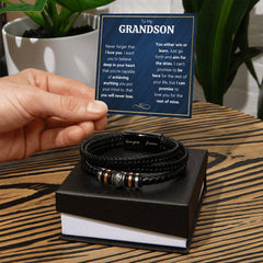 Hand holding a heartfelt message card above a braided black leather bracelet with metal accents, displayed on a black gift box with plants in the background.