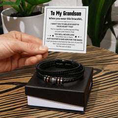 Hand holding a message card with an inspirational note for a grandson above a braided black leather bracelet with metal accents, displayed on a black gift box on a wooden surface.