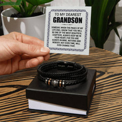 Hand holding a heartfelt message card for a grandson above a black braided leather bracelet with engraved metal accents, displayed on a black gift box on a wooden table.