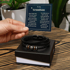 Hand holding a card with a heartfelt message "To my Grandson" beside a black braided leather bracelet with engraved metal accents, displayed on a black gift box on a wooden surface.