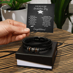 Hand holding a congratulatory card for a graduation gift next to a black braided leather bracelet with metal accents, displayed on a black gift box on a wooden surface.