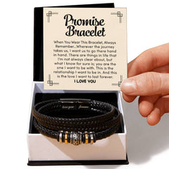 Hand holding a card with a heartfelt promise message inside an open gift box containing a black braided leather bracelet with metal accents, symbolizing love and commitment.