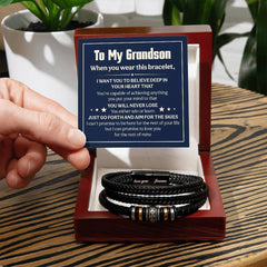 Hand holding an inspirational message card above an open wooden box containing a braided black leather bracelet with engraved metal accents, displayed on a wooden table beside potted plants.