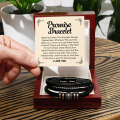 Hand holding a card with a heartfelt promise message beside a braided black leather bracelet with metallic accents, displayed in an open wooden gift box on a wooden table.