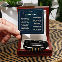 Hand holding a heartfelt message card addressed to a grandson, displayed above a black braided engraved bracelet with metal accents inside an open wooden gift box on a wooden table.